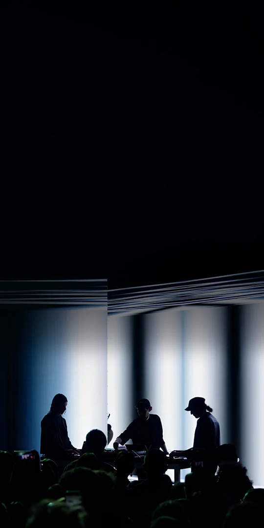 Three artists and audience silhouettes on a brightly lit stage.