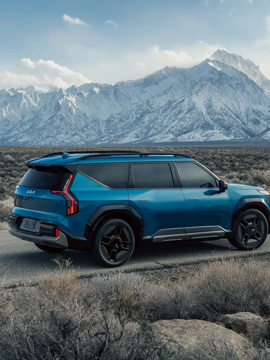 Side view of the Kia EV9 driving on a rugged road with snow-covered mountains in the background