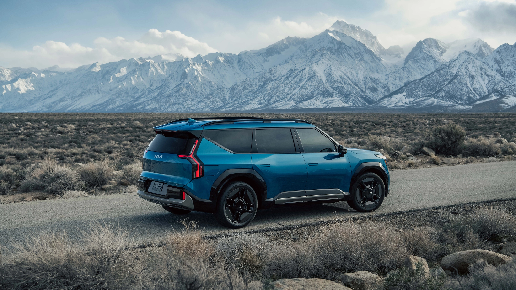 Side view of the Kia EV9 driving on a rugged road with snow-covered mountains in the background