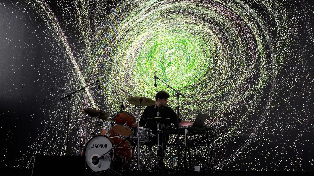 Live performance with drums and electronic equipment in front of particle-based visuals.