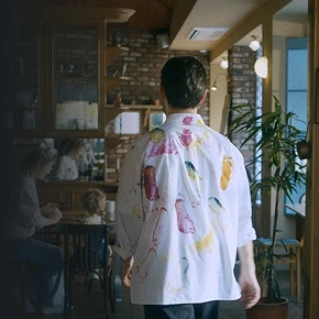 페인트가 묻은 셔츠를 입은 인물이 카페 안을 걷는 뒷모습