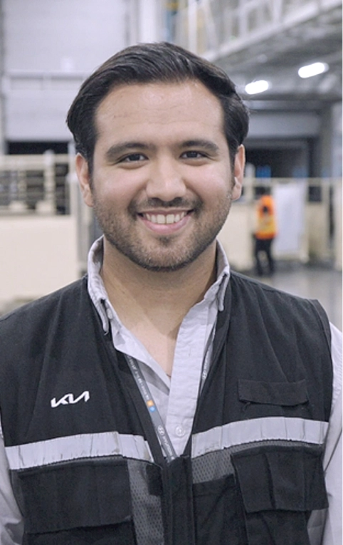 Kia Mexico employee Cesar smiling during an interview on the plant floor, wearing a KIA-branded work vest